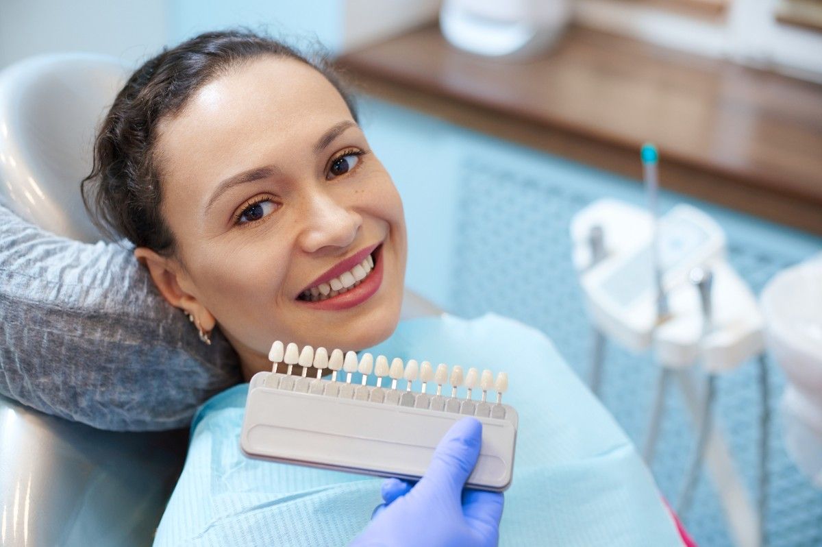brunette-woman-with-beautiful-smile-before-receiving-dental-care-check-up-teeth-whitening-bleaching-female-doctor-wearing-exam-gloves-checks-tooth-color-with-comparison-veneer-scale-chart (1)